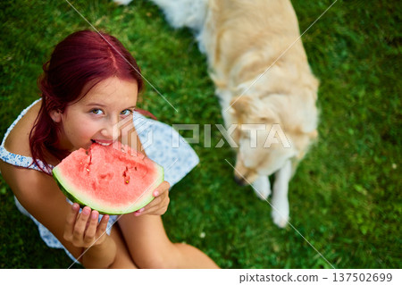 Teen girl enjoying watermelon while sitting on grass with a dog nearby during a sunny day in the backyard Teen girl enjoying watermelon while sitting on grass with a dog nearby during a sunny day in the backyard 137502699