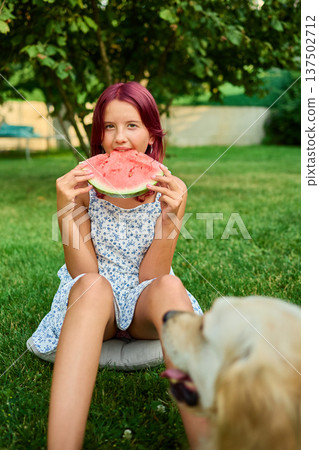 Teen girl enjoying watermelon while sitting on grass with a dog nearby during a sunny day in the backyard Teen girl enjoying watermelon while sitting on grass with a dog nearby during a sunny day in the backyard 137502712