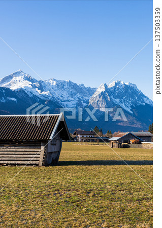 Traditional wooden hut in Bavarian countryside with snow capped Alps mountains and clear blue sky 137503359