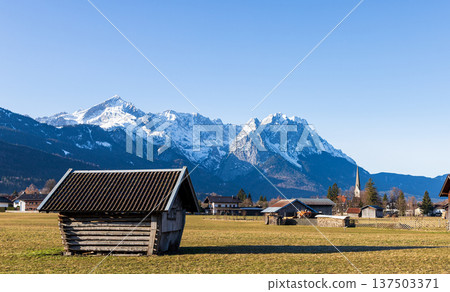 Traditional wooden hut in Bavarian countryside with snow capped Alps mountains and clear blue sky 137503371