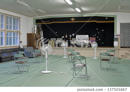 一間充滿懷舊氣息的木製學校體育館，舞台周圍擺放著折疊椅和風扇。 137503867
