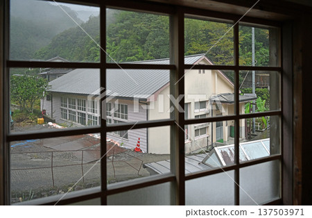 A wooden school gymnasium nestled in a misty mountain village seen through a window on a rainy day 137503971