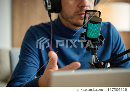 Man speaking into podcast microphone with headphones at desk 137504516