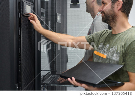 IT technician adjusting server settings in a data center rack while holding a laptop and smiling IT technician adjusting server settings in a data center rack while holding a laptop and smiling 137505418