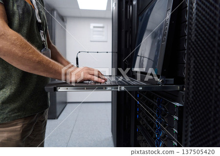 It technician working on a laptop console in a data center server rack performing network maintenance and monitoring 137505420