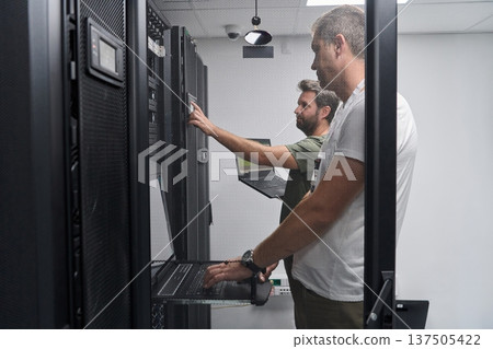 Two IT technicians troubleshooting servers in a secure data center server room with laptops 137505422