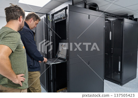 Two IT technicians working on a server rack with a laptop in a modern data center Two IT technicians working on a server rack with a laptop in a modern data center 137505423