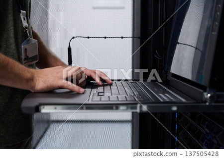 Data center technician typing on a laptop console while monitoring rack servers during system maintenance 137505428