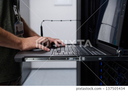 Data center technician typing on rack laptop while monitoring servers in a server room 137505434