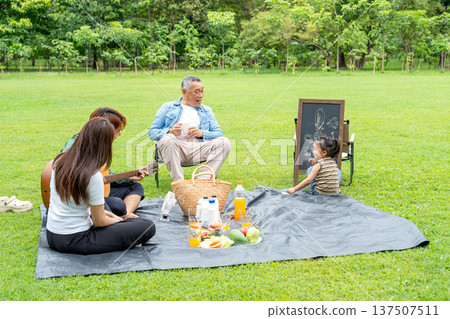 The Asian little girl turn her head to smile to grandfather after finish her drawing work and support by father and mother that they sit on mat in public park. 137507511