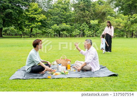 Father and grandfather sit on mat in grassfield of public park and talking together while mother and grandchild enjoy to play with daylight. 137507554