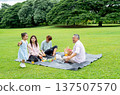 Asian little girl run around her mother, father and grandfather that they sit on mat and picnic together in public park with daylight and happiness. 137507570