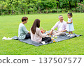 The Asian little girl stand beside grandfather and smile while her mother and father help to prepare fruit and drink for picnic in public park together with daylight. 137507577