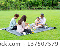 The Asian little girl sit on grandfather lap while her mother and father help to prepare fruit and drink for picnic in public park together with daylight. 137507579