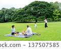 Wide shot of father and grandfather sit on mat in grassfield of public park and look mother and grandchild enjoy to play together with daylight. 137507585