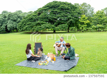 Wide shot of Asian little girl concentrate to draw and stay with father and mother also grandfather play the music using guitar and sit beside of them in public park with daylight. Wide shot of Asian little girl concentrate to draw and stay with father and mother also grandfather play the music using guitar and sit beside of them in public park with daylight. 137507591
