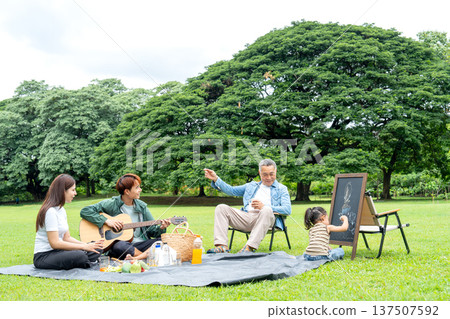 Side view of Asian grandfather sit on chair and look at grandchild draw while young man play music using guitar in public park during they have holiday together with daylight. Side view of Asian grandfather sit on chair and look at grandchild draw while young man play music using guitar in public park during they have holiday together with daylight. 137507592