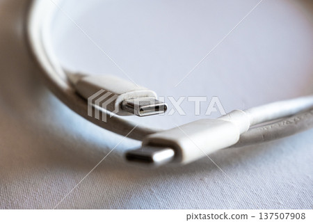 Close-up of a type-C cable with two connectors. The cable is white and flexible, lying on a light surface. Ideal for technology and electronics themes. Close-up of a type-C cable with two connectors. The cable is white and flexible, lying on a light surface. Ideal for technology and electronics themes. 137507908