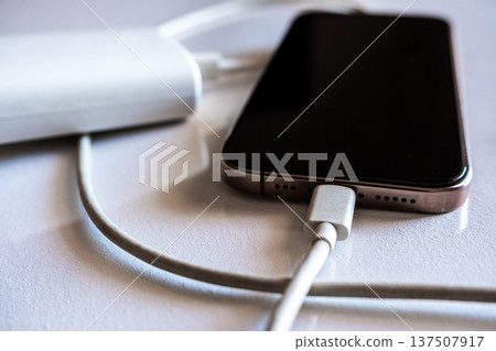 A smartphone is charging with a power bank. The phone is silver with a black screen. The power bank is white and connected with a charging cable. A smartphone is charging with a power bank. The phone is silver with a black screen. The power bank is white and connected with a charging cable. 137507917