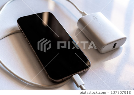 A smartphone is charging with a power bank. The phone is silver with a black screen. The power bank is white and connected with a charging cable. A smartphone is charging with a power bank. The phone is silver with a black screen. The power bank is white and connected with a charging cable. 137507918