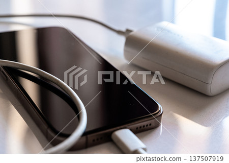 A smartphone is charging with a power bank. The phone is silver with a black screen. The power bank is white and connected with a charging cable. A smartphone is charging with a power bank. The phone is silver with a black screen. The power bank is white and connected with a charging cable. 137507919
