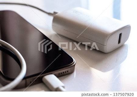 A smartphone is charging with a power bank. The phone is silver with a black screen. The power bank is white and connected with a charging cable. A smartphone is charging with a power bank. The phone is silver with a black screen. The power bank is white and connected with a charging cable. 137507920