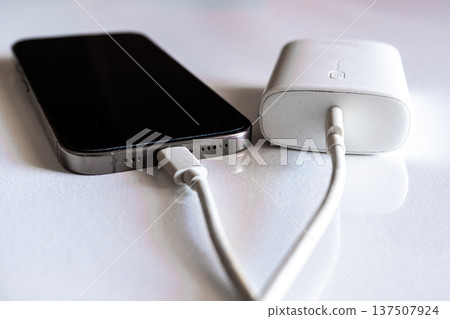 A smartphone is charging with a power bank. The phone is silver with a black screen. The power bank is white and connected with a charging cable. A smartphone is charging with a power bank. The phone is silver with a black screen. The power bank is white and connected with a charging cable. 137507924