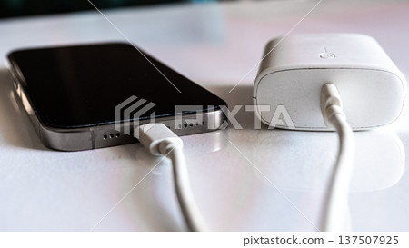 A smartphone is charging with a power bank. The phone is silver with a black screen. The power bank is white and connected with a charging cable. A smartphone is charging with a power bank. The phone is silver with a black screen. The power bank is white and connected with a charging cable. 137507925