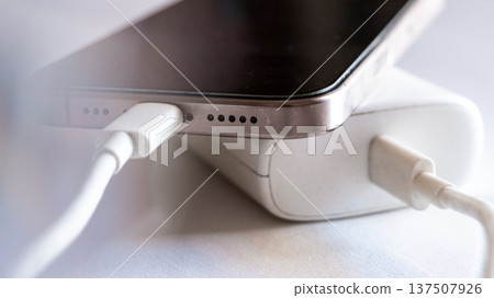 A smartphone is charging with a power bank. The phone is silver with a black screen. The power bank is white and connected with a charging cable. A smartphone is charging with a power bank. The phone is silver with a black screen. The power bank is white and connected with a charging cable. 137507926