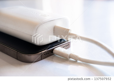 A smartphone is charging with a power bank. The phone is silver with a black screen. The power bank is white and connected with a charging cable. A smartphone is charging with a power bank. The phone is silver with a black screen. The power bank is white and connected with a charging cable. 137507932