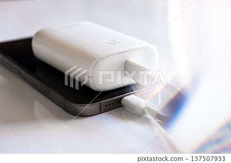 A smartphone is charging with a power bank. The phone is silver with a black screen. The power bank is white and connected with a charging cable. 137507933