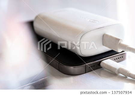 A smartphone is charging with a power bank. The phone is silver with a black screen. The power bank is white and connected with a charging cable. A smartphone is charging with a power bank. The phone is silver with a black screen. The power bank is white and connected with a charging cable. 137507934