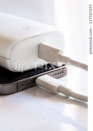 A smartphone is charging with a power bank. The phone is silver with a black screen. The power bank is white and connected with a charging cable. A smartphone is charging with a power bank. The phone is silver with a black screen. The power bank is white and connected with a charging cable. 137507935