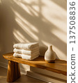 Morning light across spa bench, folded towels sit beside diffuser, soft shadows on wall, towels stacked neatly on warm wood, towels suggest cozy wellness, minimalist decor vibe, calm home bath theme 137508836