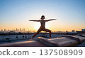 Woman practices yoga on rooftop sunrise. Warrior pose against city skyline, yoga inspiration for International Yoga Day, summer solstice wellness ads, copy space, mindful lifestyle concept 137508909