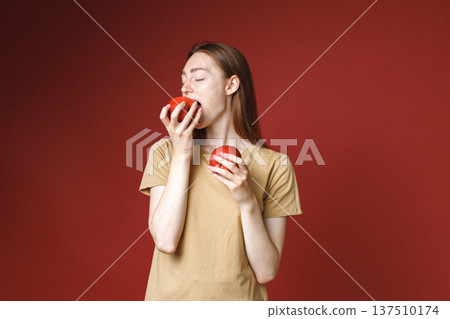 Studio portrait of caucasian girl isolated on red background. Portrait of young beautiful woman in beige t-shirt posing for a photo. Studio photo of a young girl holding two red apples. Studio portrait of caucasian girl isolated on red background. Portrait of young beautiful woman in beige t-shirt posing for a photo. Studio photo of a young girl holding two red apples. 137510174