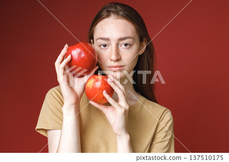 Studio portrait of caucasian girl isolated on red background. Portrait of young beautiful woman in beige t-shirt posing for a photo. Studio photo of a young girl holding two red apples. Studio portrait of caucasian girl isolated on red background. Portrait of young beautiful woman in beige t-shirt posing for a photo. Studio photo of a young girl holding two red apples. 137510175