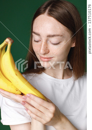 Indoor portrait of caucasian girl isolated on green background. Portrait of young beautiful woman in white t-shirt cheerfuly posing for a photo. Studio photo of a young girl holding three bananas. Indoor portrait of caucasian girl isolated on green background. Portrait of young beautiful woman in white t-shirt cheerfuly posing for a photo. Studio photo of a young girl holding three bananas. 137510176