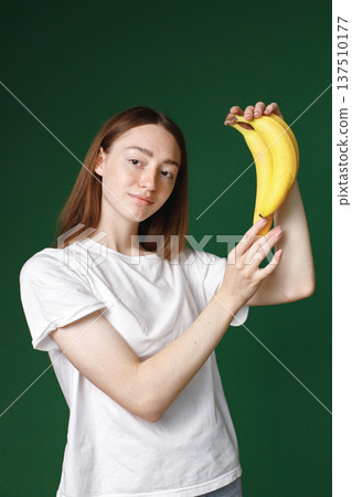 Indoor portrait of caucasian girl isolated on green background. Portrait of young beautiful woman in white t-shirt cheerfuly posing for a photo. Studio photo of a young girl holding three bananas. Indoor portrait of caucasian girl isolated on green background. Portrait of young beautiful woman in white t-shirt cheerfuly posing for a photo. Studio photo of a young girl holding three bananas. 137510177