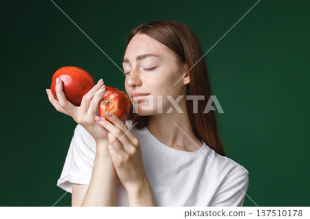 Indoor portrait of caucasian girl isolated on green background. Portrait of young beautiful woman in white t-shirt cheerfuly posing for a photo. Studio photo of a young girl holding two red apples. Indoor portrait of caucasian girl isolated on green background. Portrait of young beautiful woman in white t-shirt cheerfuly posing for a photo. Studio photo of a young girl holding two red apples. 137510178