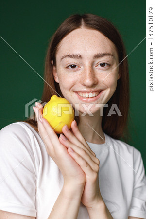 Indoor portrait of caucasian girl isolated on green background. Portrait of young beautiful woman in white t-shirt cheerfuly posing for a photo. Studio photo of a young girl holding a lemon. 137510179