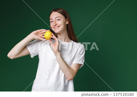 Indoor portrait of caucasian girl isolated on green background. Portrait of young beautiful woman in white t-shirt posing for a photo. Studio photo of a young girl holding a lemon. 137510180