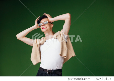 Young blond man with make up on his face and sunglasses. Man wearing fashionable white t-shirt and black cap. Man isolated on green background. Young blond man with make up on his face and sunglasses. Man wearing fashionable white t-shirt and black cap. Man isolated on green background. 137510197