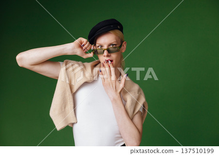 Young blond man with make up on his face and sunglasses. Man wearing fashionable white t-shirt and black cap. Man isolated on green background. Young blond man with make up on his face and sunglasses. Man wearing fashionable white t-shirt and black cap. Man isolated on green background. 137510199