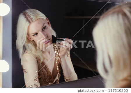 Young blond man holding brushes and doing make up on his face. Man wearing fashionable bright golden t-shirt. Man looking in a studio mirror. Young blond man holding brushes and doing make up on his face. Man wearing fashionable bright golden t-shirt. Man looking in a studio mirror. 137510200