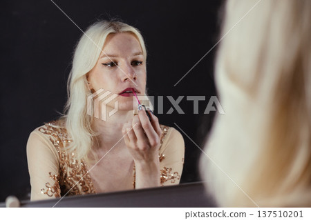 Young blond man holding brushes and doing make up on his face. Man wearing fashionable bright golden t-shirt. Man looking in a studio mirror. 137510201