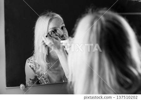 Young blond man holding brushes and doing make up on his face. Man wearing fashionable t-shirt. Man looking in a studio mirror. Young blond man holding brushes and doing make up on his face. Man wearing fashionable t-shirt. Man looking in a studio mirror. 137510202
