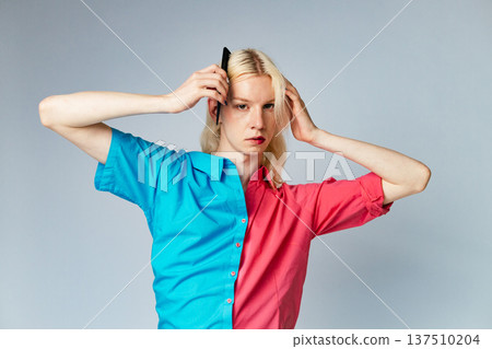 Young blond man has make up on one half of his face. Man wearing fashionable shirt divided on two colours bright pink and blue. Man isolated on grey background. 137510204