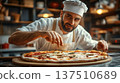 Male chef in white uniform and hat, skillfully garnishing freshly made pizza with herbs and spices, showcasing culinary artistry in a rustic kitchen environment 137510689