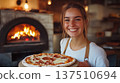 Young woman with brown hair, smiling while holding freshly baked pizza in a rustic kitchen, with a wood-fired oven glowing in the background, showcasing culinary passion and warmth 137510694